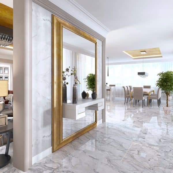 Intérieur moderne avec sols en marbre, grand miroir encadré d'or, table console décorative, et vue sur une salle à manger et un salon avec lumière naturelle et plantes.