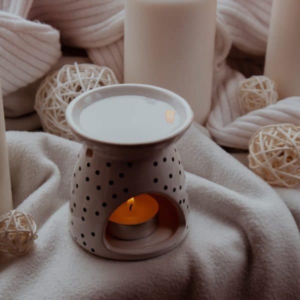 Une bougie allumée se trouve à l'intérieur d'un support en céramique avec un motif à pois, entouré de bougies blanches et de boules décoratives en osier sur un fond en tissu doux.