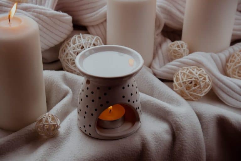 Une bougie allumée se trouve à l'intérieur d'un support en céramique avec un motif à pois, entouré de bougies blanches et de boules décoratives en osier sur un fond en tissu doux.