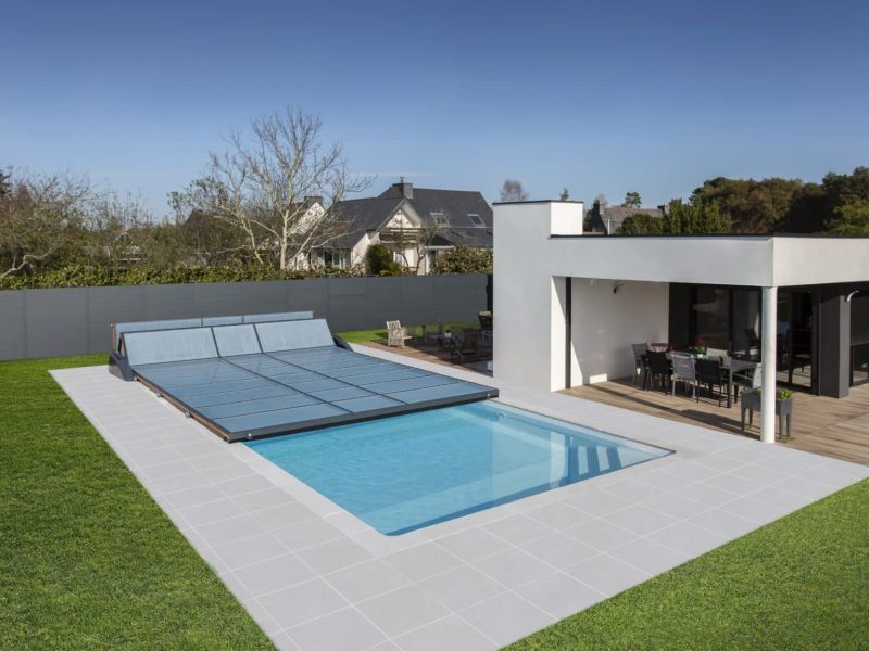 Une cour arrière moderne avec une piscine extérieure rectangulaire, partiellement couverte par une couverture de piscine, à côté d'un bâtiment de patio blanc avec des sièges extérieurs.
