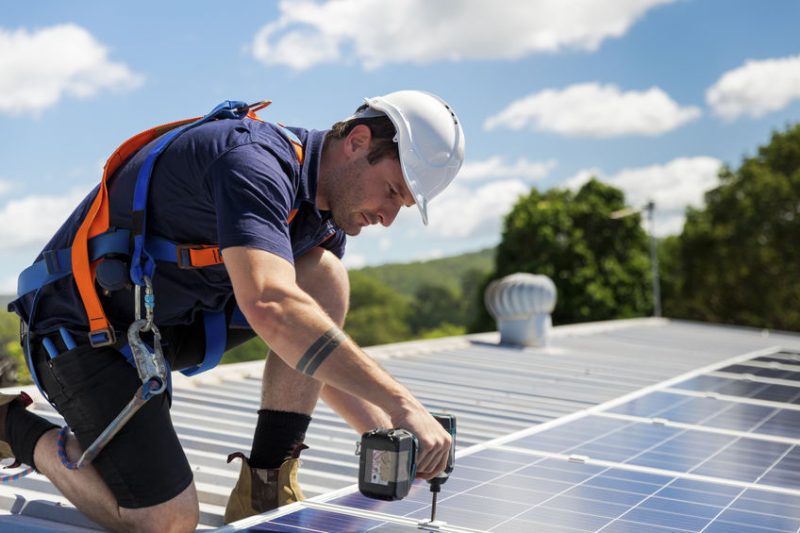 Un ouvrier portant un équipement de sécurité et un casque utilise une perceuse électrique pour installer des panneaux solaires sur un toit sous un ciel partiellement nuageux.