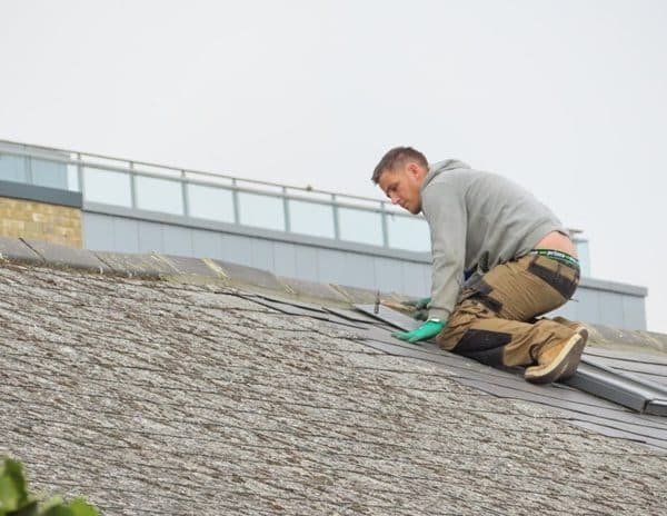 Un homme portant un sweat à capuche gris et un pantalon marron travaille sur un toit en pente et ajuste les bardeaux. Un bâtiment est visible en arrière plan.