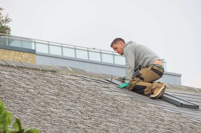 Un homme portant un sweat à capuche gris et un pantalon marron travaille sur un toit en pente et ajuste les bardeaux. Un bâtiment est visible en arrière plan.