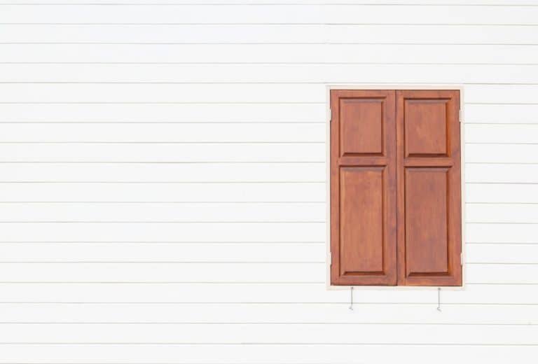 Un volet de fenêtre en bois marron fermé sur un mur de parement horizontal blanc.
