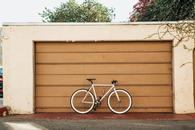 Un vélo blanc avec un guidon et un siège noirs s'appuie contre une porte de garage fermée et marron clair avec des plantes qui poussent autour.