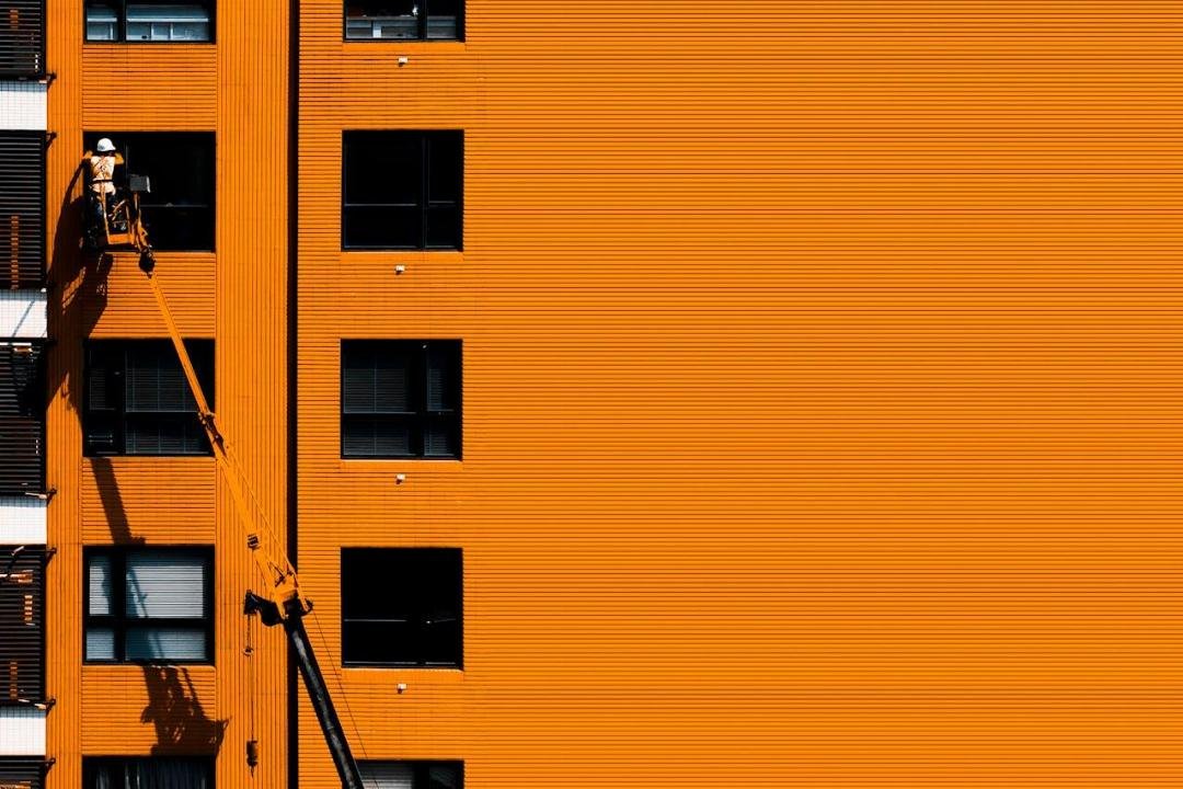 Un ouvrier dans un ascenseur inspecte ou répare la façade orange d'un bâtiment doté de plusieurs fenêtres régulièrement espacées.