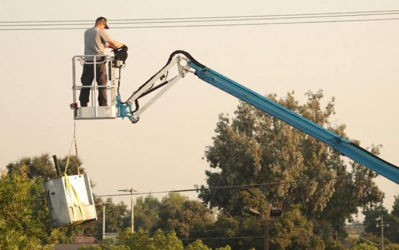 Un homme exploite une nacelle bleue et hisse un gros objet métallique dans un décor extérieur avec des arbres et des lignes électriques en arrière plan.