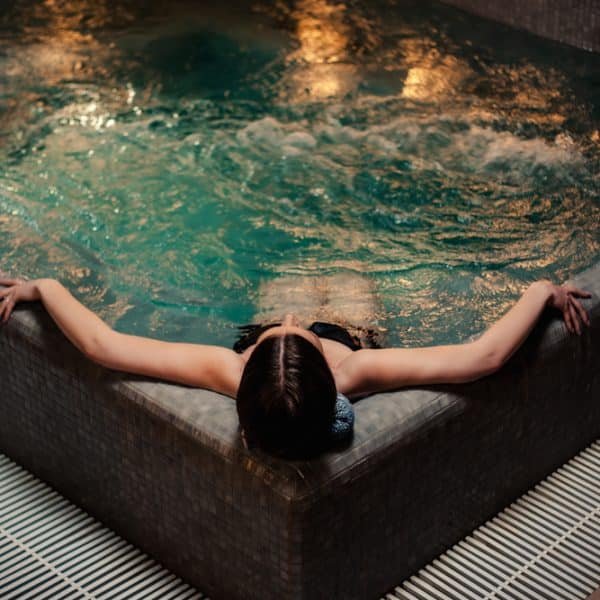 Une personne vêtue d'un maillot de bain noir se détend, les bras tendus, sur le bord d'un jacuzzi ou d'un bain à remous intérieur, entouré d'un sol carrelé.