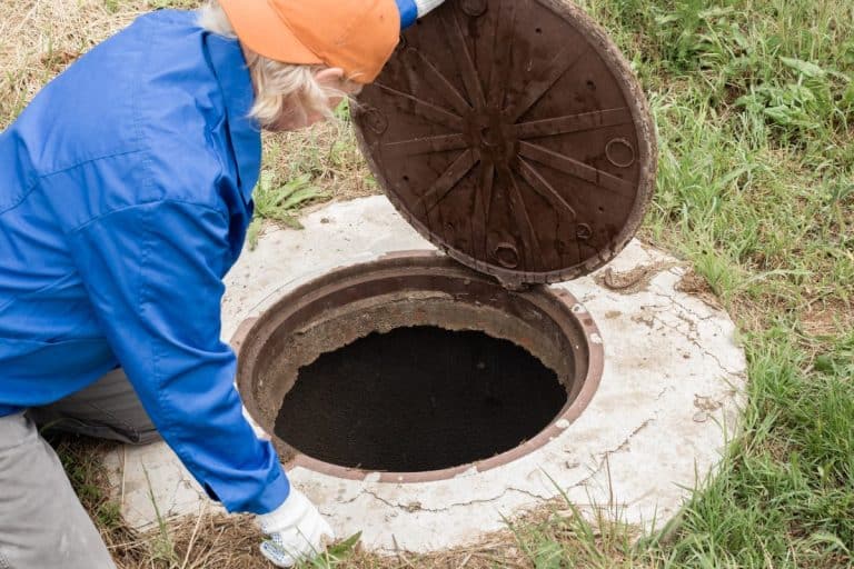Une personne agenouillée soulève un couvercle de regard rond en métal dans une zone herbeuse.