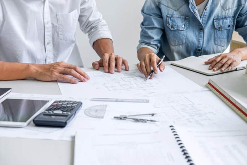 Deux personnes discutent de plans architecturaux avec des outils comme une calculatrice, des stylos et des cahiers sur la table.