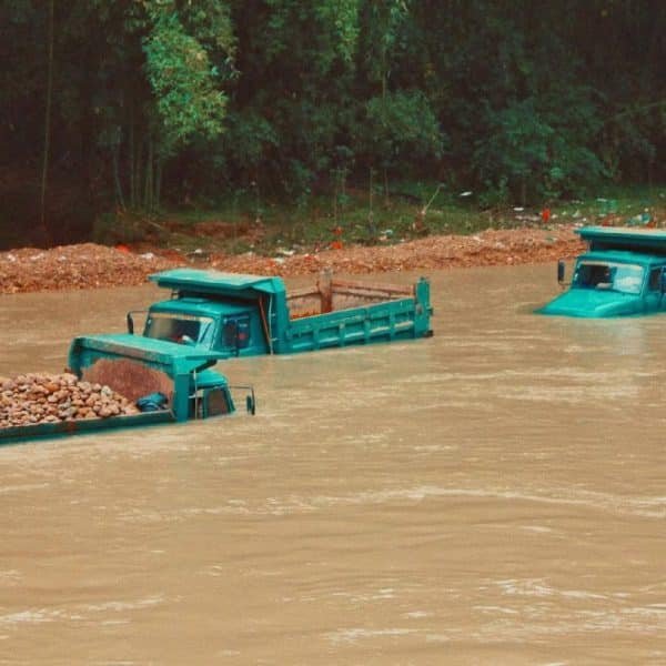 Deux camions bleus immergés dans une rivière en crue transportant des charges de roches, avec un talus boueux en arrière plan.