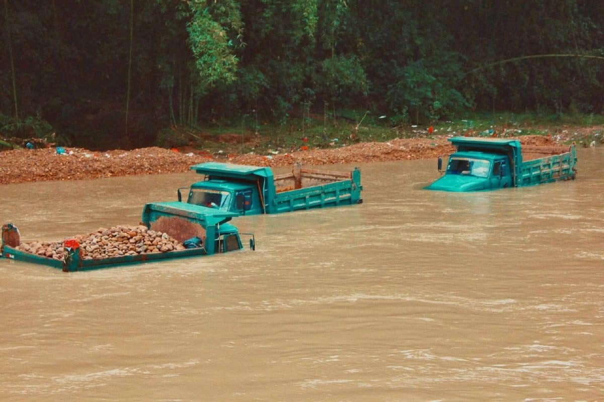 Deux camions bleus immergés dans une rivière en crue transportant des charges de roches, avec un talus boueux en arrière plan.