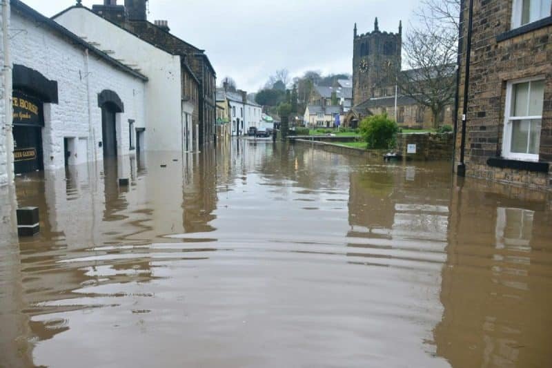 Une rue inondée dans une ville avec des routes submergées à proximité de bâtiments traditionnels en pierre et une église en arrière plan par temps couvert.