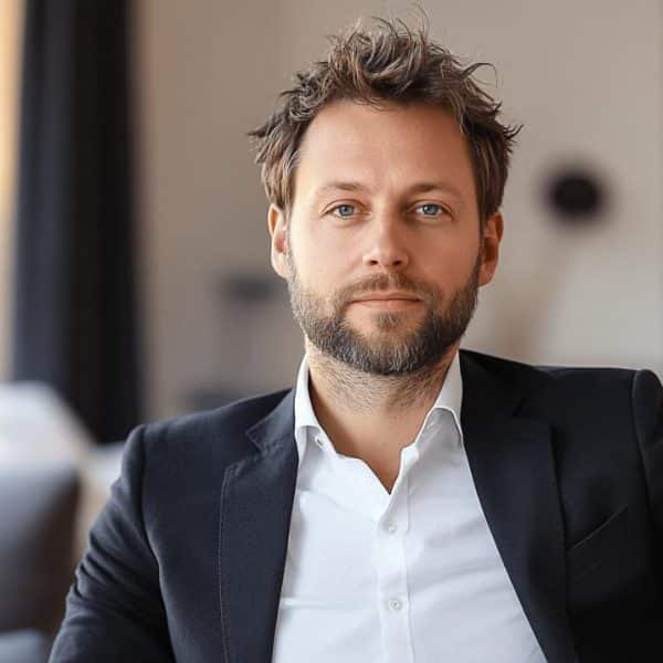 Un homme vêtu d'un blazer noir et d'une chemise blanche est assis dans une pièce bien éclairée avec des éléments d'arrière plan flous.