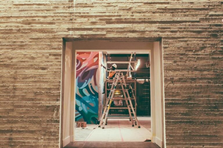 Une personne se tient sur une échelle pour peindre une peinture murale colorée sur le mur d'un bâtiment, vue à travers une porte dans un mur de béton texturé.