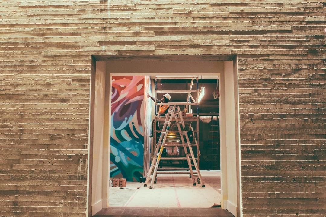 Une personne se tient sur une échelle pour peindre une peinture murale colorée sur le mur d'un bâtiment, vue à travers une porte dans un mur de béton texturé.