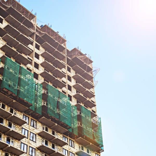 Un immeuble de grande hauteur en construction avec des échafaudages et des filets de sécurité verts sur la façade contre un ciel clair.