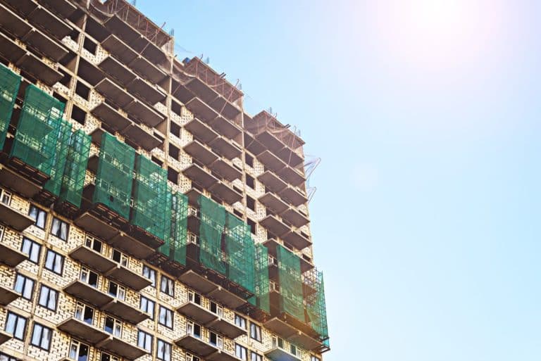 Un immeuble de grande hauteur en construction avec des échafaudages et des filets de sécurité verts sur la façade contre un ciel clair.