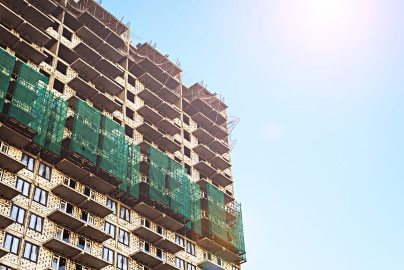 Un immeuble de grande hauteur en construction avec des échafaudages et des filets de sécurité verts sur la façade contre un ciel clair.