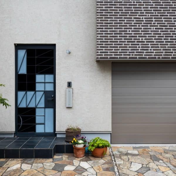 Entrée de maison moderne avec une porte à motifs géométriques, une porte de garage adjacente, des plantes en pot sur un chemin carrelé et une façade en briques.