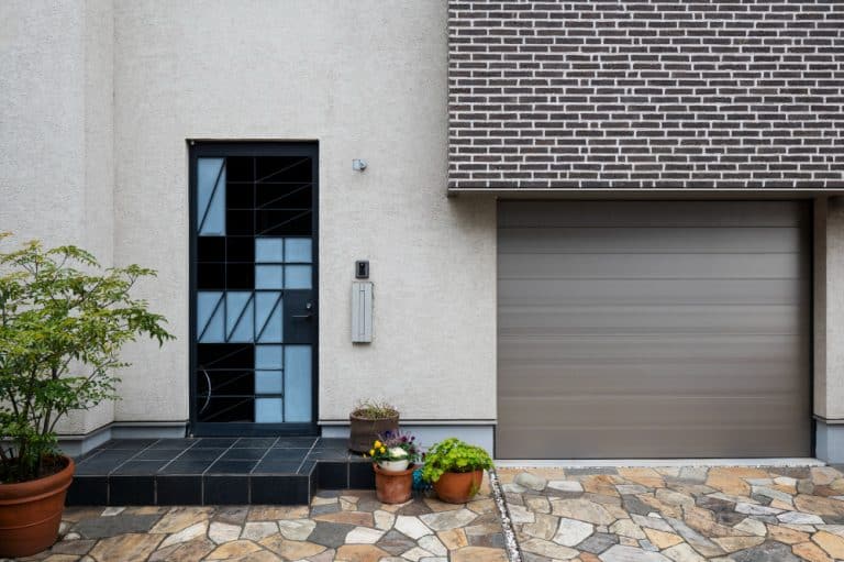 Entrée de maison moderne avec une porte à motifs géométriques, une porte de garage adjacente, des plantes en pot sur un chemin carrelé et une façade en briques.