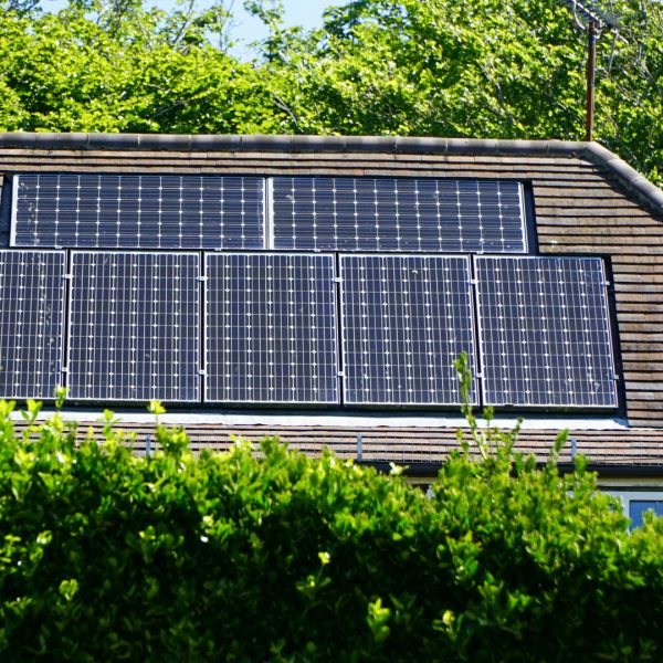Plusieurs panneaux solaires sont installés sur le toit incliné d'une maison, partiellement obscurci par des buissons verts, avec des arbres et un ciel bleu en arrière plan.