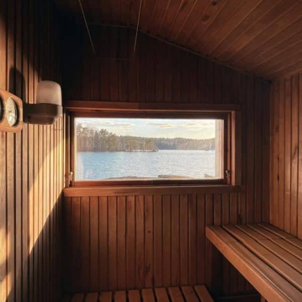 Un intérieur de sauna en bois avec une fenêtre offrant une vue sur un lac serein et un rivage boisé. Des bancs sont placés le long des murs et des instruments de sauna sont montés à côté de la fenêtre.