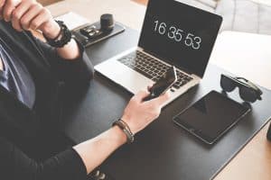 Personne à un bureau tenant un smartphone, un ordinateur portable affichant l’heure, une tablette et des lunettes de soleil placées à proximité.
