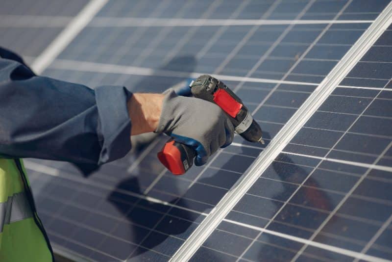 Une personne portant des gants utilise une perceuse électrique pour installer ou entretenir un panneau solaire à l'extérieur.