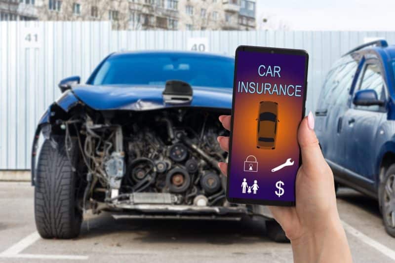 Une main tient un smartphone affichant une application d'assurance automobile devant une voiture endommagée avec l'avant écrasé sur un parking. Une main tient un smartphone affichant une application d'assurance automobile devant une voiture endommagée avec l'avant écrasé sur un parking.
