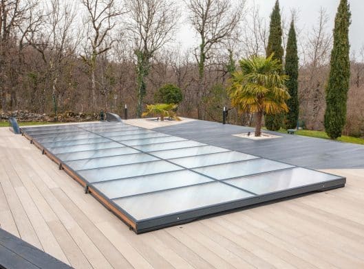 Une piscine extérieure rectangulaire est recouverte d'une couverture de piscine transparente et plate sur une terrasse en bois entourée d'arbres et de plantes paysagères.