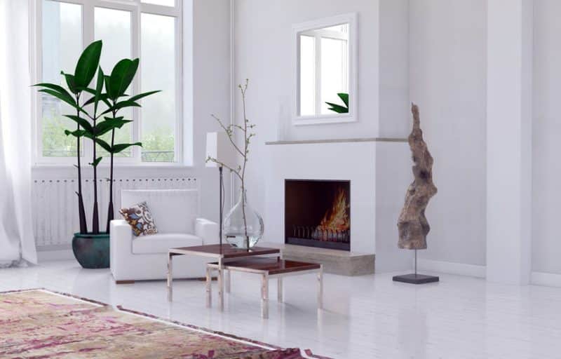 Salon moderne avec des murs blancs, une cheminée, une grande plante verte, un fauteuil blanc, des vases en verre sur une table basse, une sculpture abstraite et une grande fenêtre laissant entrer la lumière naturelle.