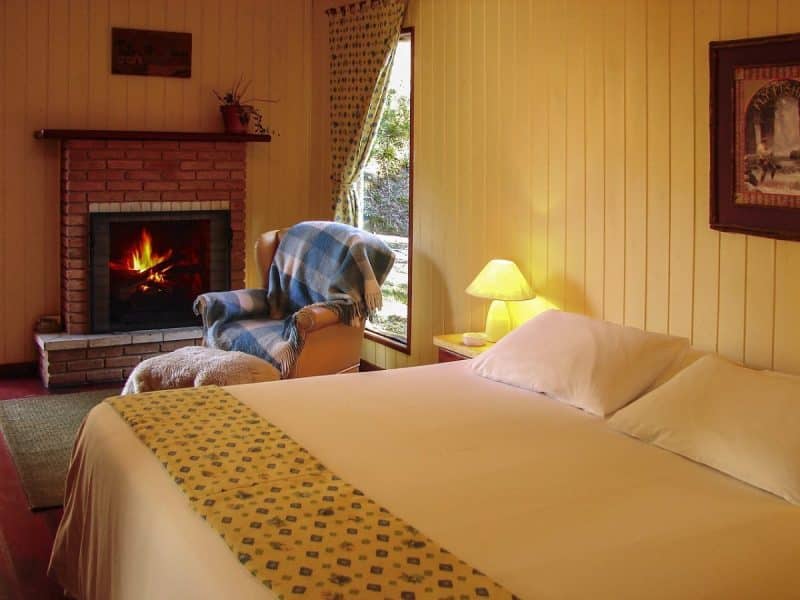Chambre cosy avec cheminée, fauteuil et lit fait. Une fenêtre avec rideau drapé laisse entrer la lumière naturelle. Une lampe et un tableau encadré complètent le décor.