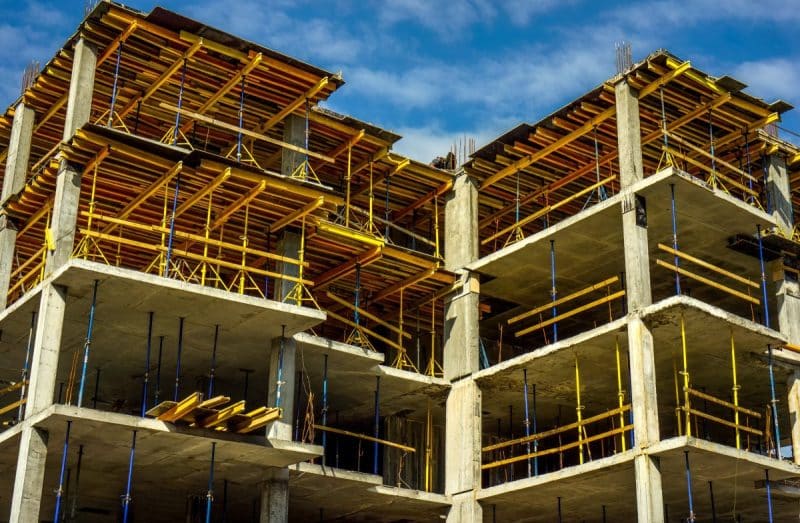 Un bâtiment à plusieurs étages en construction avec des sols en béton, des supports verticaux et des échafaudages jaunes sur un ciel bleu.