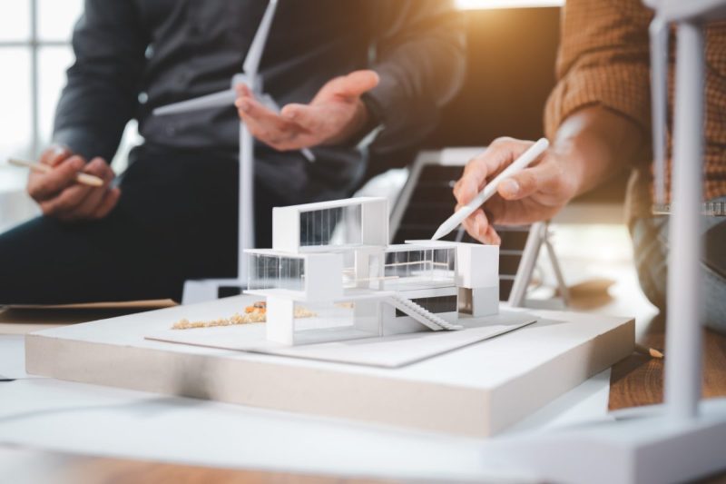 Deux personnes discutent et font un geste vers un modèle architectural moderne d'un bâtiment sur un bureau, avec des matériaux et des outils de conception à proximité.