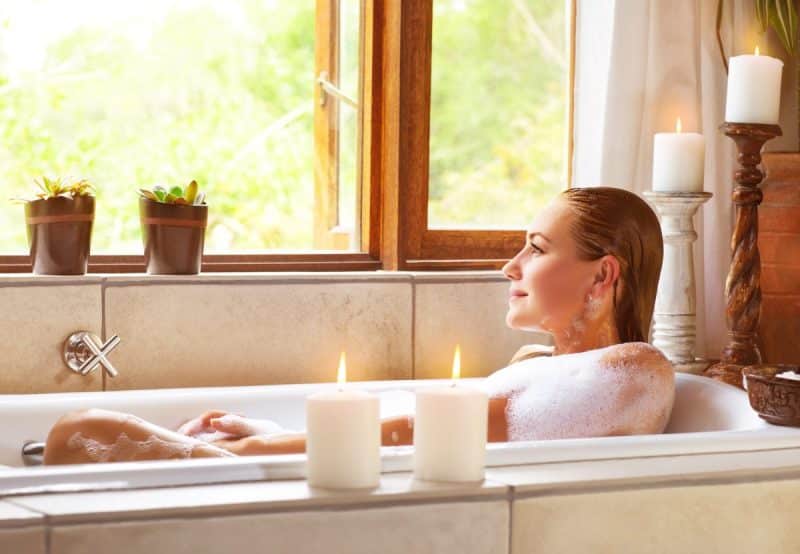 Une personne se détend dans une baignoire remplie de bulles près d’une fenêtre avec des plantes vertes et des bougies allumées sur le rebord.
