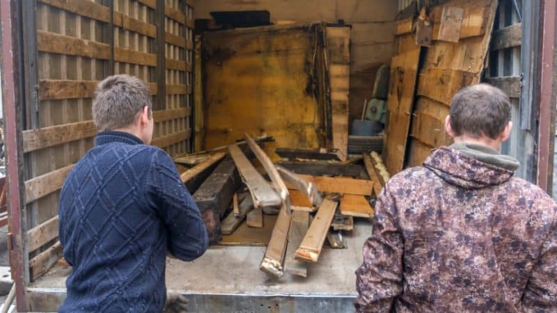 Deux hommes en tenue décontractée chargent ou déchargent des débris de bois de l'arrière d'un camion. Le camion semble partiellement rempli de divers morceaux de bois et de matériaux de construction.