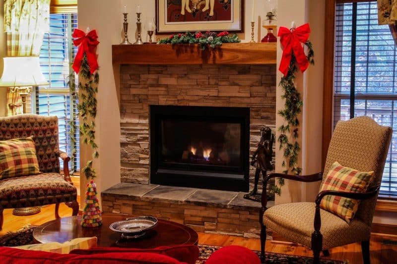 Salon chaleureux avec cheminée en pierre flanquée de guirlandes festives et de noeuds rouges. Deux fauteuils entourent une table basse avec un arbre décoratif. De grandes fenêtres avec stores apportent de la lumière naturelle.