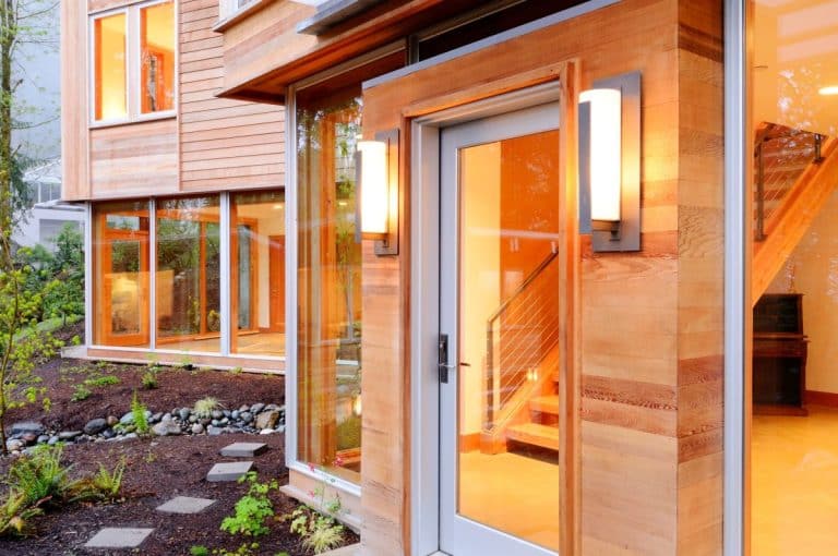 Extérieur de maison moderne avec de grandes fenêtres et un revêtement en bois. L'entrée éclairée est dotée d'une porte vitrée encadrée de deux appliques. Des marches et des plantes vertes ornent le jardin paysager.
