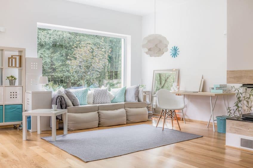 Salon moderne avec grande fenêtre, canapé beige, coussins assortis, tapis gris, chaise blanche et suspension. Bureau et étagères avec décor. Verdure visible à l'extérieur. Salon moderne avec grande fenêtre, canapé beige, coussins assortis, tapis gris, chaise blanche et suspension. Bureau et étagères avec décor. Verdure visible à l'extérieur.