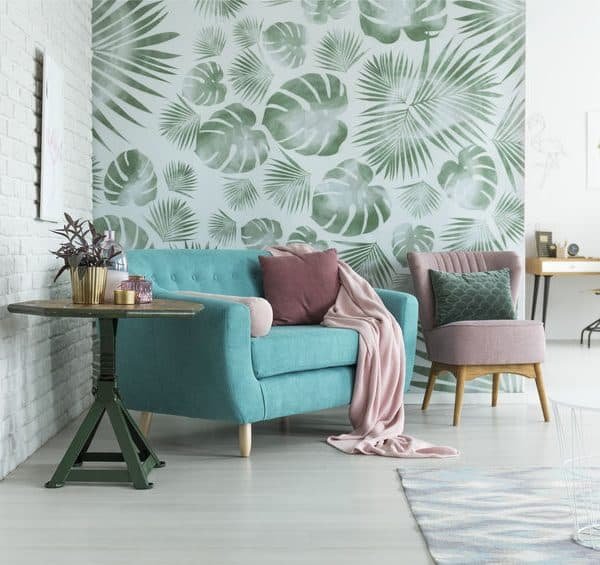 Salon avec un canapé bleu sarcelle, des coussins roses et verts, une chaise rose, du papier peint à feuilles, une table ronde avec des plantes et un bureau en arrière plan.