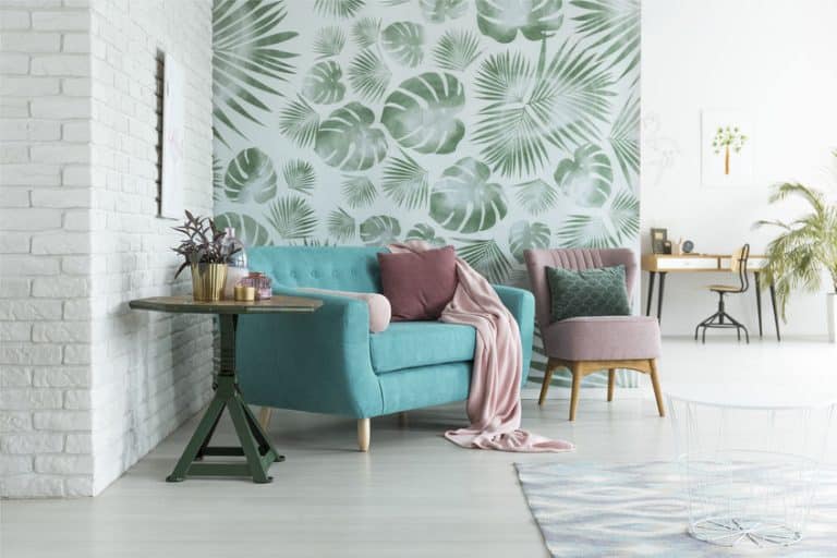 Salon avec un canapé bleu sarcelle, des coussins roses et verts, une chaise rose, du papier peint à feuilles, une table ronde avec des plantes et un bureau en arrière plan.