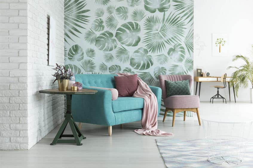 Salon avec un canapé bleu sarcelle, des coussins roses et verts, une chaise rose, du papier peint à feuilles, une table ronde avec des plantes et un bureau en arrière plan.