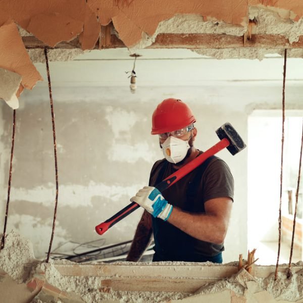 Un ouvrier du bâtiment portant un casque de sécurité rouge, un masque et des gants tient une masse tout en démolissant un mur intérieur.