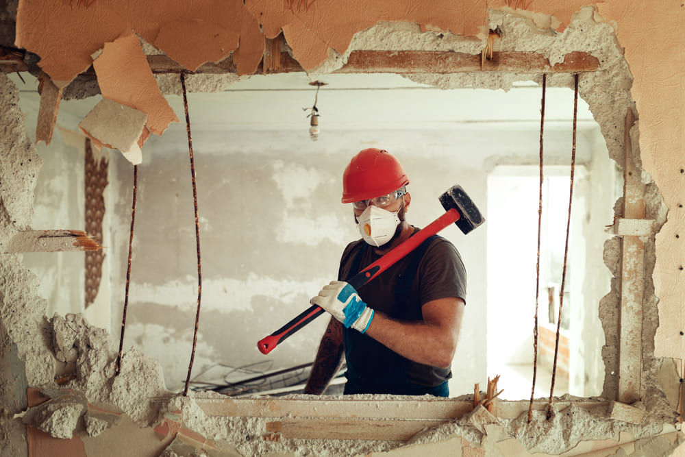 Un ouvrier du bâtiment portant un casque de sécurité rouge, un masque et des gants tient une masse tout en démolissant un mur intérieur.