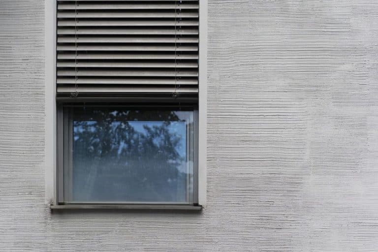 Une fenêtre fermée avec des stores horizontaux partiellement tirés sur la moitié supérieure est insérée dans le mur extérieur gris texturé d'un bâtiment.
