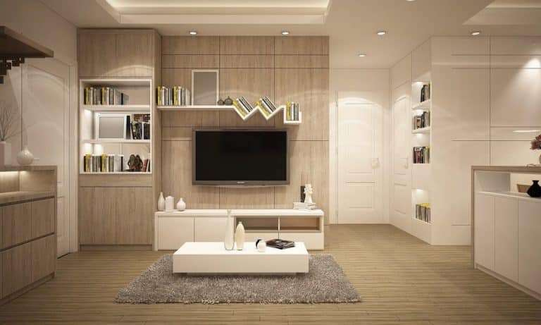 Salon moderne avec une télévision murale, des étagères avec des livres, des touches de bois, une table basse blanche et un tapis gris. Des plafonniers illuminent l'espace.