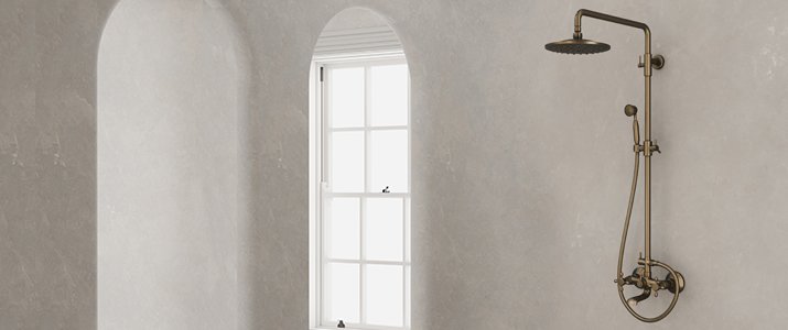 Une robinetterie de douche en bronze est fixée sur un mur gris clair, près d'une fenêtre cintrée aux encadrements blancs, qui laisse entrer la lumière naturelle.