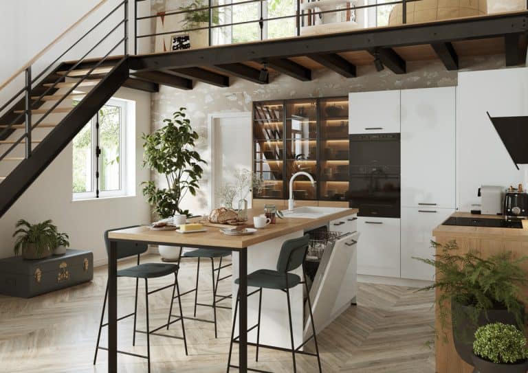Cuisine moderne avec armoires blanches, îlot avec comptoir en bois, tabourets de bar, appareils encastrés, plantes et étagères ouvertes sous un escalier dans un espace de style loft.