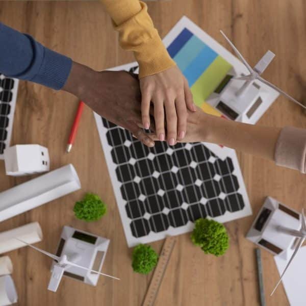Trois personnes se tiennent la main au dessus d'une table sur laquelle se trouvent des schémas de panneaux solaires, des maquettes d'éoliennes et des plans d'architecture, suggérant un travail d'équipe dans le cadre d'un projet d'énergie renouvelable.